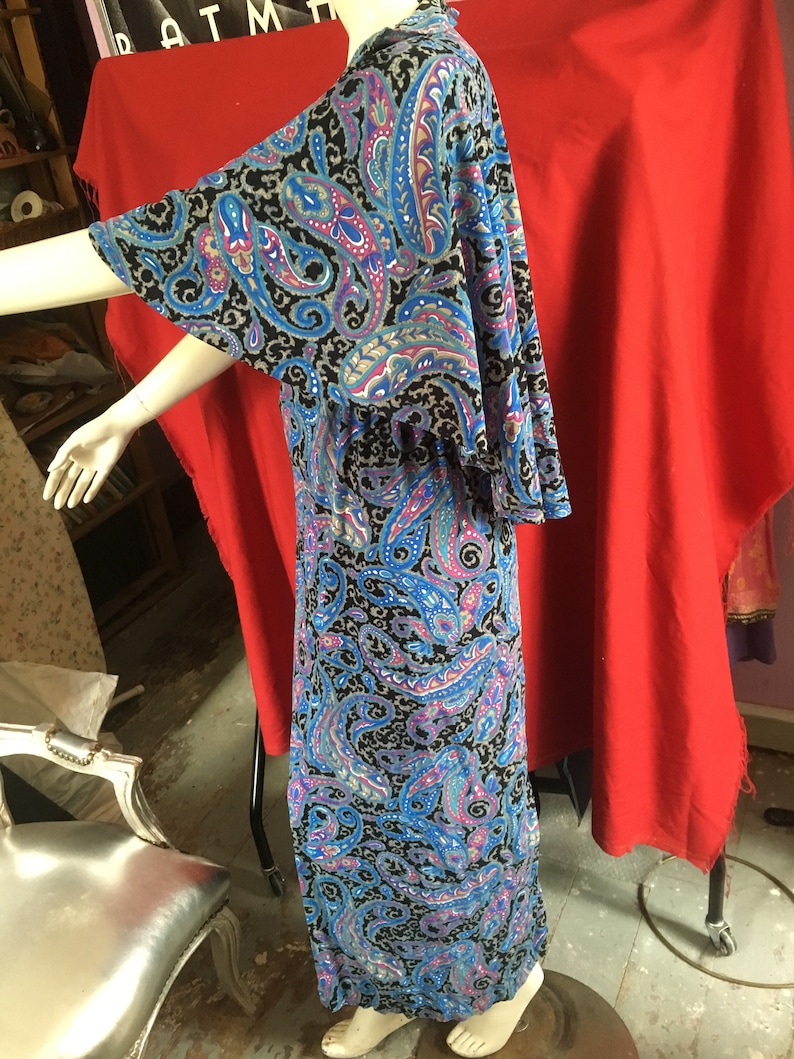 May include: A long, flowing maxi dress with a paisley print in shades of blue, pink, and gray on a black background. The dress has a wide, flowing neckline and bell sleeves.