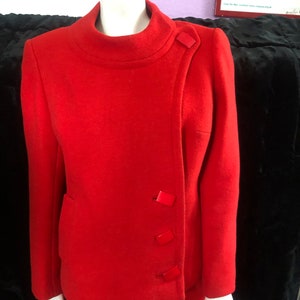 K&ouml;nnte beinhalten: Ein roter Wollanzug mit Stehkragen und Rock. Die Jacke hat einen schr&auml;gen Verschluss mit vier roten Kn&ouml;pfen.
