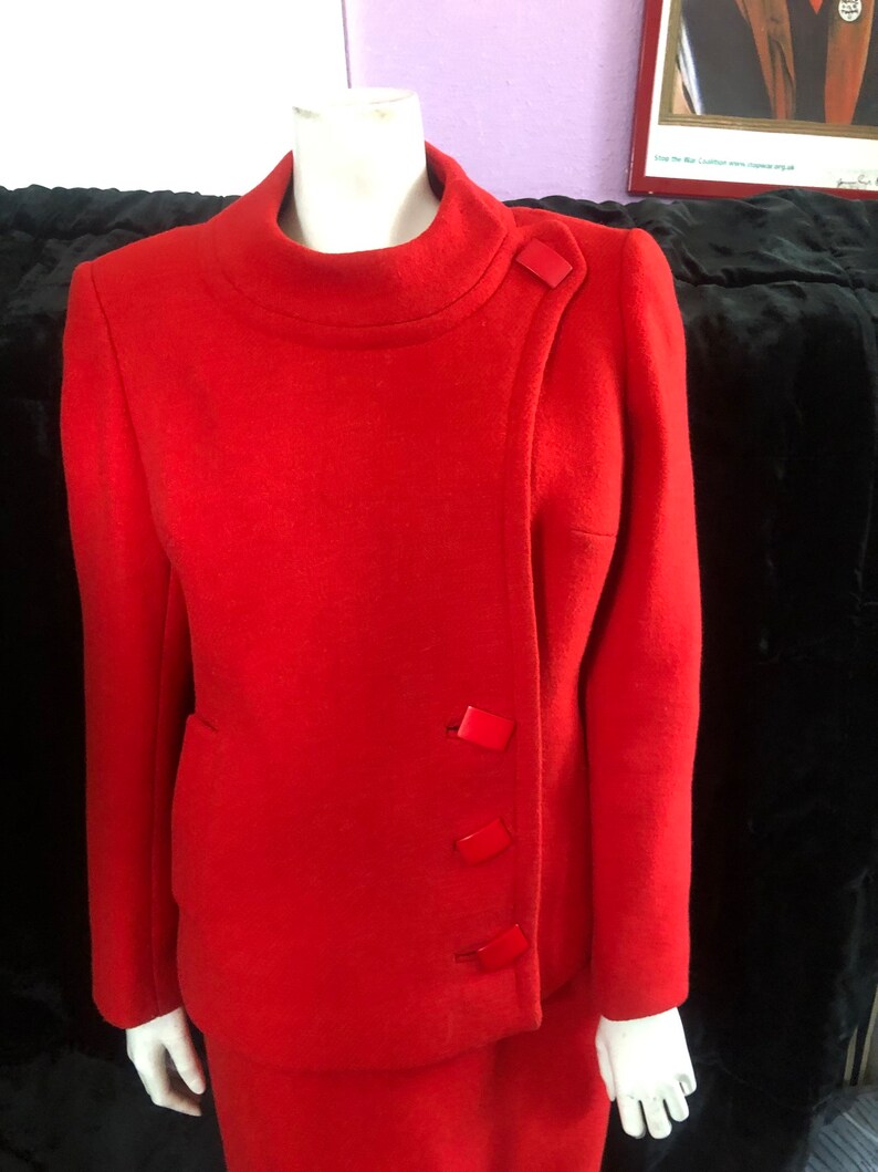 K&ouml;nnte beinhalten: Ein roter Wollanzug mit Stehkragen und Rock. Die Jacke hat einen schr&auml;gen Verschluss mit vier roten Kn&ouml;pfen.