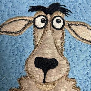 Könnte beinhalten: Ein braunes und schwarzes gesticktes Tier mit großen Augen und einer angenähten Nase. Das Tier befindet sich auf einem blauen Hintergrund mit einem gesteppten Muster.