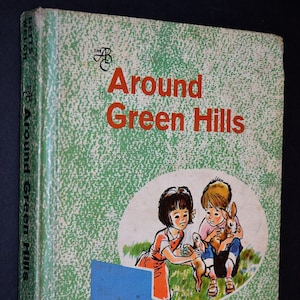 May include: Vintage children's book titled "Around Green Hills." The cover features illustrations of two children with rabbits, set against a green speckled background. The title is in orange, with a blue square and orange circle.