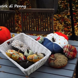 Peut inclure: Un panier en osier blanc rempli de citrouilles en tissu colorées. Les citrouilles sont décorées de différents motifs et tissus, notamment du denim, du plaid et des imprimés floraux. Le panier est posé sur une table grise avec une vraie citrouille orange et des feuilles d'automne éparpillées autour.
