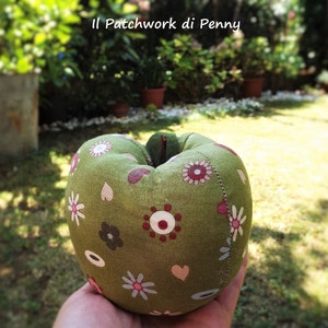 May include: A green fabric apple with a patchwork design featuring flowers, hearts, and circles in shades of red, white, and brown. The apple is sitting on a hand.