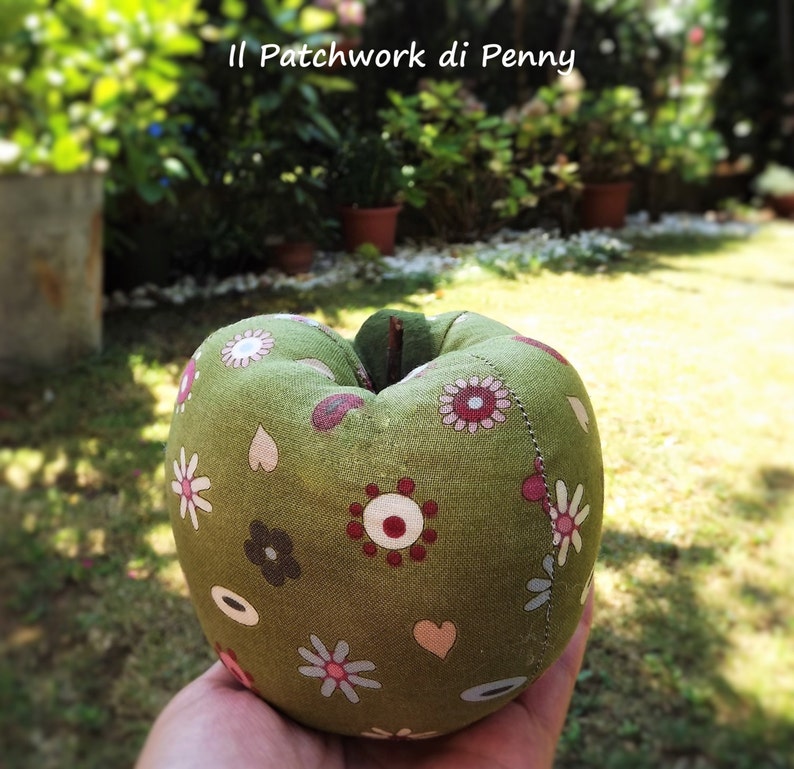 May include: A green fabric apple with a patchwork design featuring flowers, hearts, and circles in shades of red, white, and brown. The apple is sitting on a hand.