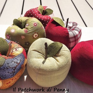 Op de afbeelding: Zes handgemaakte stoffen appelnaaldenkussens in verschillende kleuren en patronen, waaronder rood, groen en wit. De naaldenkussens zijn in een halve cirkel op een wit oppervlak gerangschikt. De tekst "Il Patchwork di Penny" is zichtbaar onderaan de afbeelding.