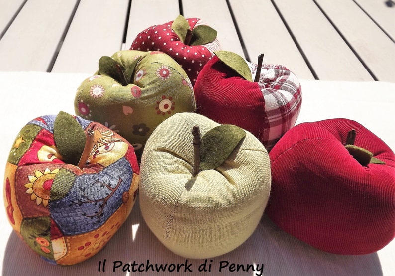 May include: Six handmade fabric apple pincushions in various colors and patterns, including red, green, and white. The pincushions are arranged in a semi-circle on a white surface. The text "Il Patchwork di Penny" is visible at the bottom of the image.
