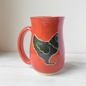 May include: A ceramic mug with a bright red glaze and a black chicken silhouette. The mug has a curved shape with a handle and a cream-colored base. This kitchenware item is suitable for hot or cold beverages.