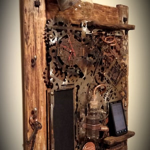 Puede incluir: Una estantería de pared de madera rústica con un reloj, engranajes y otros elementos de inspiración steampunk. La estantería tiene una pequeña pizarra, una lámpara y un soporte para teléfono.