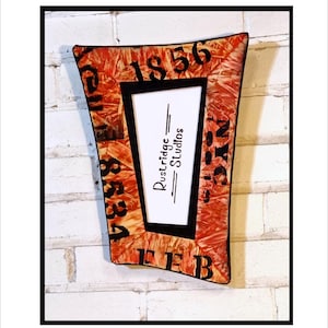 May include: A decorative, trapezoid-shaped picture frame with a black border. The frame has a reddish-orange, textured surface with black lettering and the text "Rustridge Studios". The frame is mounted on a white brick wall.