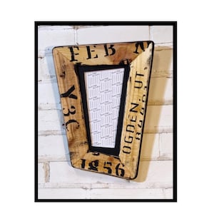 May include: Wooden picture frame with a black border. The frame has a rustic, distressed look with black lettering and numbers, including "FEB", "Y3C", "OGDEN UT", and "1956". The frame holds a white and gray patterned image.