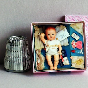 May include: A miniature doll with light skin and molded hair, wearing a white outfit, presented in a square box with accessories including tiny clothing, a bottle, and a tag. A silver-toned metal thimble is positioned next to the box.