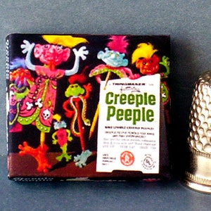 May include: A box of Creepy Peeple, a toy with a colorful illustration of a green, red, and pink creature with long legs and arms. The box has a tag that reads 'Thinkmaker Creepy Peeple, Nine Livable Creepy Peeple.'  Next to the box is a silver thimble.