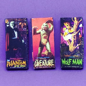 May include: Three miniature vintage horror movie boxes featuring "Phantom of the Opera", "The Creature", and "Wolf Man" on a purple surface. Each box has colorful illustrations and text.