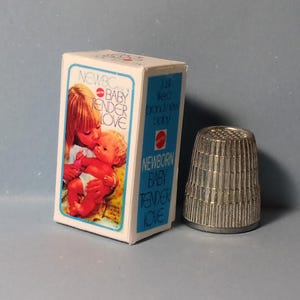May include: A vintage toy box with the words "NEWBORN BABY TENDER LOVE" and an image of a woman kissing a baby. A silver thimble is next to the box. The box is white with blue trim.