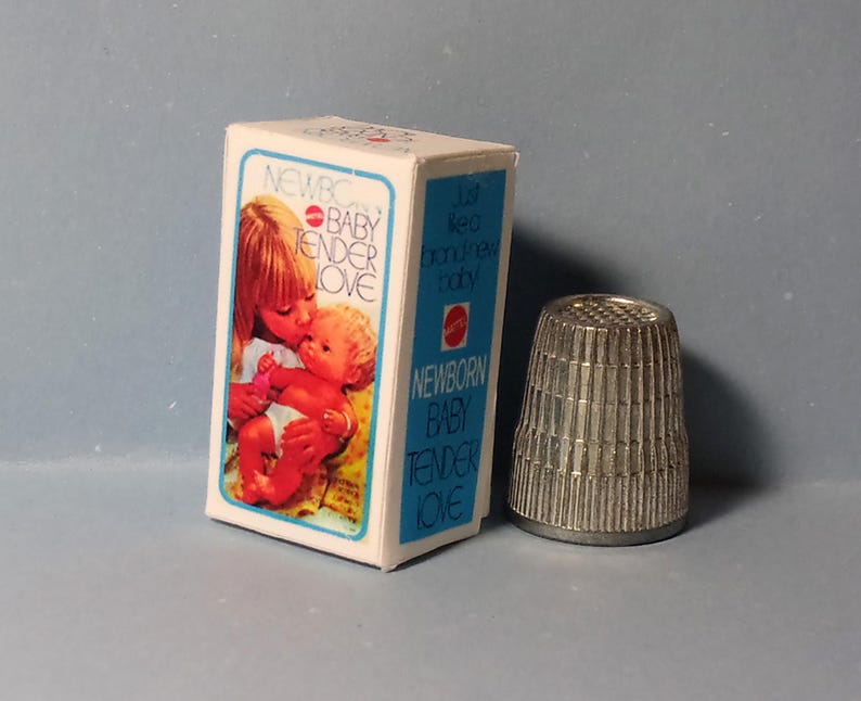 May include: A vintage toy box with the words "NEWBORN BABY TENDER LOVE" and an image of a woman kissing a baby. A silver thimble is next to the box. The box is white with blue trim.