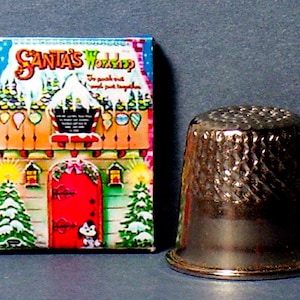 May include: A vintage Christmas-themed box with a colorful illustration of Santa's Workshop. The box has a red door and a small dog peeking out. The text on the box reads "Santa's Workshop" and "To punch out and put together". A silver thimble is also pictured.