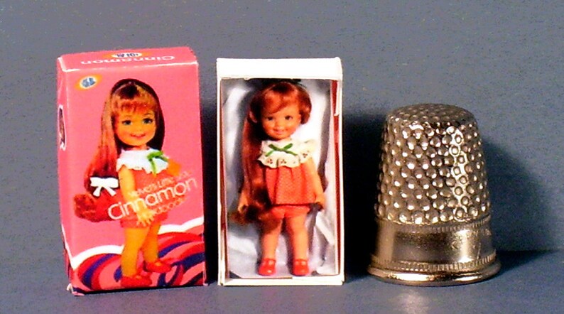 May include: A miniature doll named "Cinnamon" in a pink and white dress with a green bow. The doll is packaged in a pink box with the doll's name on it. The doll is next to a white box with the doll inside. A silver thimble is next to the boxes.