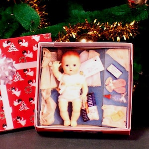 May include: A red and white gift box with a white bow and a doll inside. The doll is wearing a white dress and a white diaper. The box is sitting on a green surface.