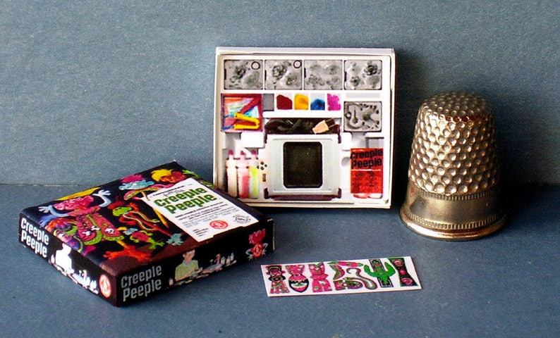 K&ouml;nnte beinhalten: Ein Vintage-Bastelset von Creepy Peeple mit einer schwarz-wei&szlig;en Schachtel mit bunten Illustrationen von Monstern und einer wei&szlig;en Schachtel mit einem Projektor, Farbe und Stempeln. Das Set enth&auml;lt auch eine Bogen mit Aufklebern in verschiedenen Monsterformen.