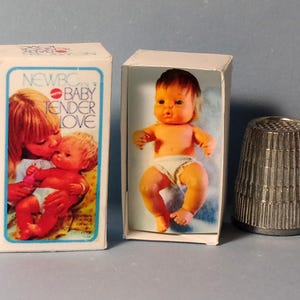 May include: Two small baby dolls in boxes with the text "NEWBC BABY TENDER LOVE" and a metal thimble. The baby dolls are orange and white. The thimble is silver.