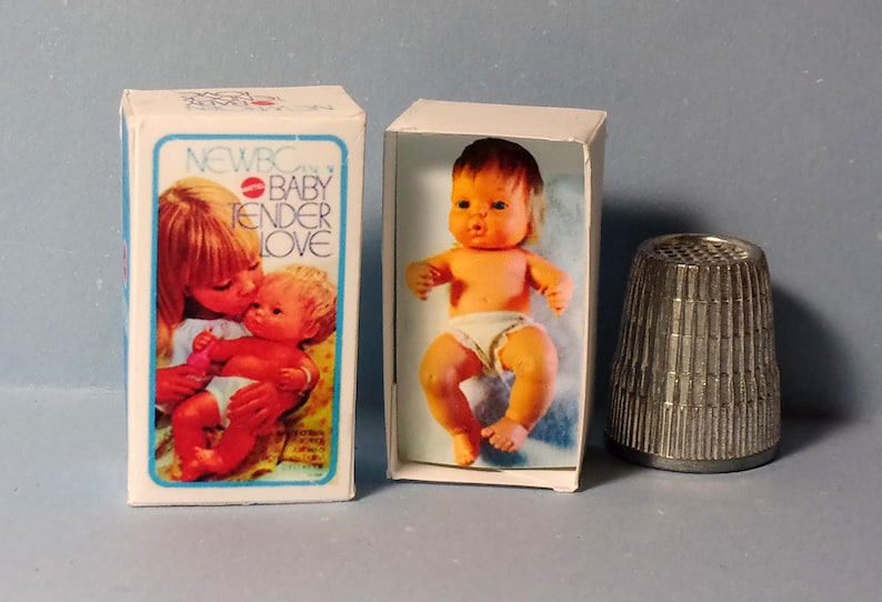 May include: Two small baby dolls in boxes with the text "NEWBC BABY TENDER LOVE" and a metal thimble. The baby dolls are orange and white. The thimble is silver.