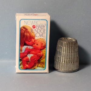 May include: A vintage "NEWBORN BABY TENDER LOVE" doll box with an illustration of a woman kissing a baby. Next to the box is a silver metal thimble with vertical grooves. The thimble is approximately 1 inch tall.