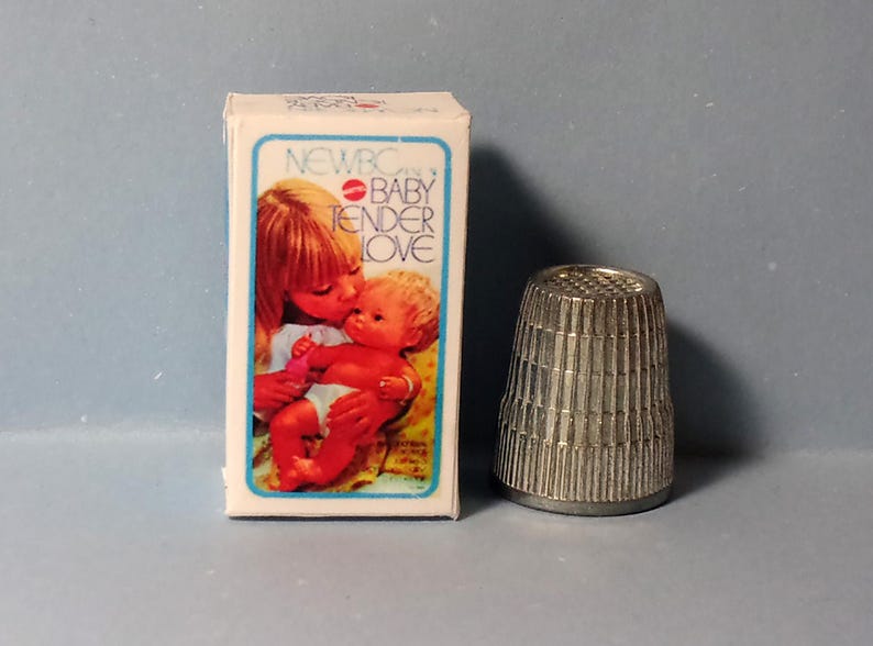 May include: A vintage "NEWBORN BABY TENDER LOVE" doll box with an illustration of a woman kissing a baby. Next to the box is a silver metal thimble with vertical grooves. The thimble is approximately 1 inch tall.