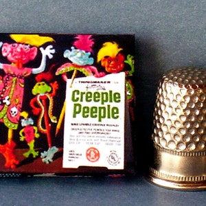K&ouml;nnte beinhalten: Eine Schachtel mit "Creeple Peeple" biegsamen gruseligen Puppen. Die Schachtel zeigt bunte Illustrationen der Puppen, darunter eine gr&uuml;ne Kreatur mit langem Hals und eine rosa Kreatur mit langem Rock. Die Schachtel steht neben einem silbernen Fingerhut.