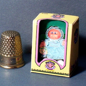 May include: A small Cabbage Patch Kids doll in a blue dress with a pink and white lace trim, holding a small plush toy. The doll is in a yellow box with a green background and the words "Cabbage Patch Kids" printed on the front.