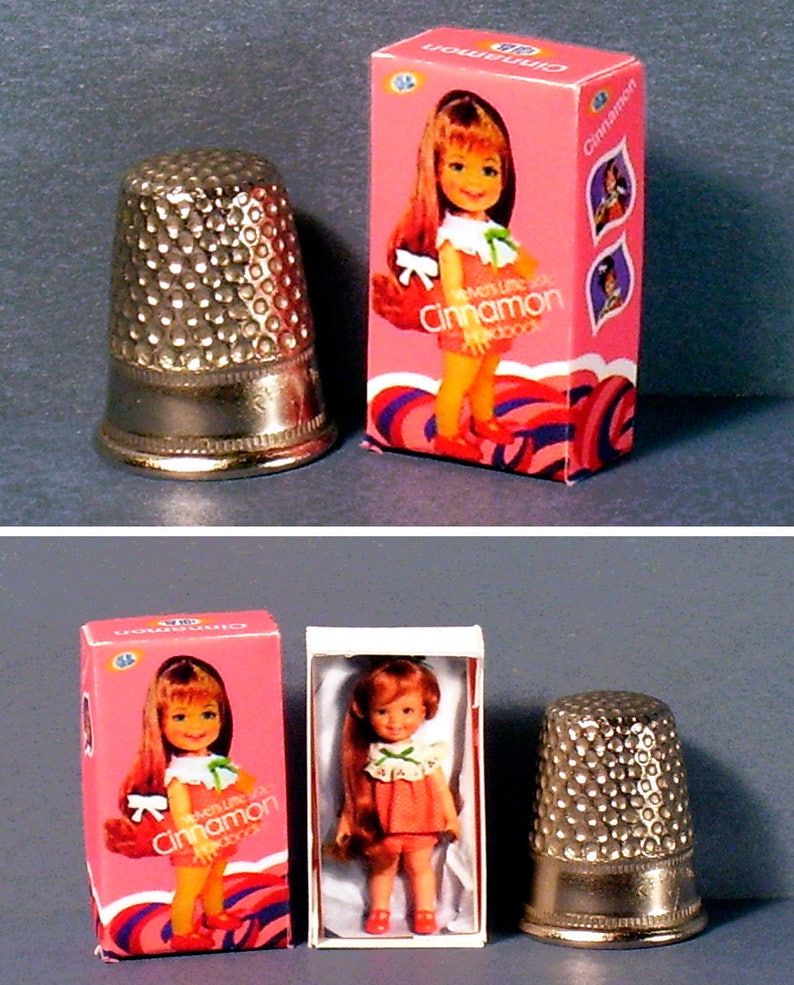 May include: A vintage doll in a pink and white box with the words "Little Miss Cinnamon" on the front. The doll is wearing a red and white dress and has long brown hair. The box is next to a silver thimble.