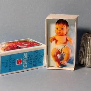 May include: A vintage doll in a box with the words "NEWBORN BABY TENDER LOVE" and a metal thimble. The doll is orange and wearing a diaper. The box is white with blue text.