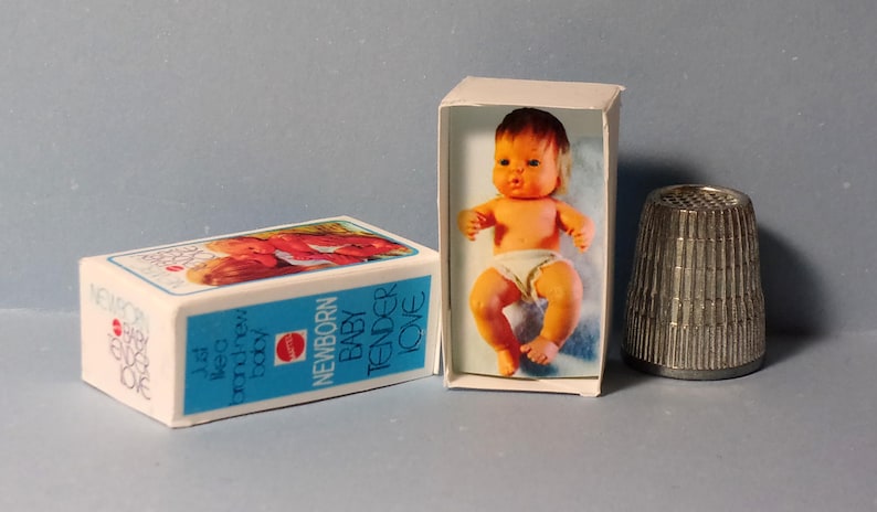 May include: A vintage doll in a box with the words "NEWBORN BABY TENDER LOVE" and a metal thimble. The doll is orange and wearing a diaper. The box is white with blue text.