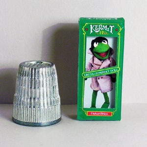 May include: A silver thimble with vertical ridges next to a green box featuring a Kermit the Frog doll in a pink coat. The box reads "Kermit the Frog" and "Dress Up Muppet Doll". The Fisher Price logo is at the bottom.