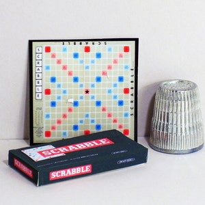 May include: A Scrabble game set with a red, white, and blue game board and a silver tile holder. The game board has the word "Scrabble" printed on it. The tile holder is a cylindrical shape with a ribbed design.
