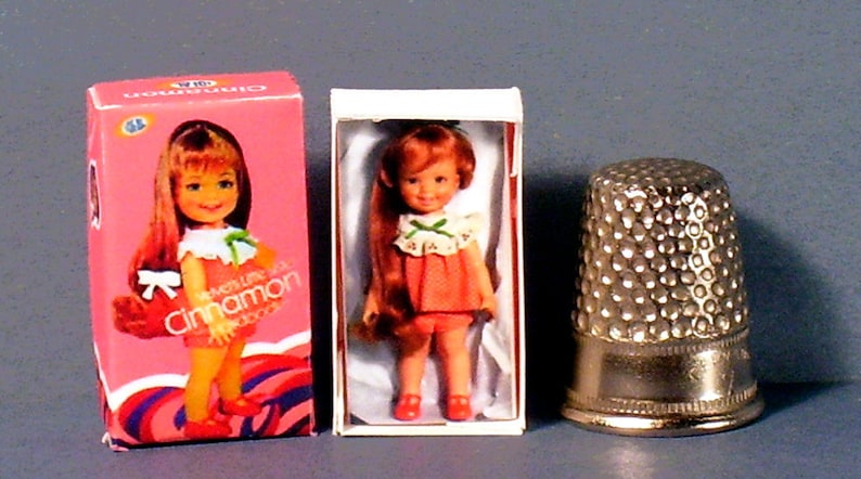 May include: A doll in a pink dress and red shoes in a white box. The doll has long brown hair and is holding a green bow. The box has the words 'The Little Miss Cinnamon Doll' printed on it. Next to the box is a silver thimble.