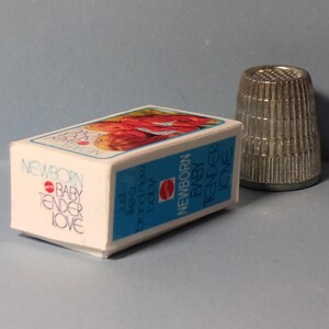 May include: A small white box with the words "NEWBORN BABY TENDER LOVE" in a script font, with a blue border and a picture of a baby on top. A silver thimble is next to the box.