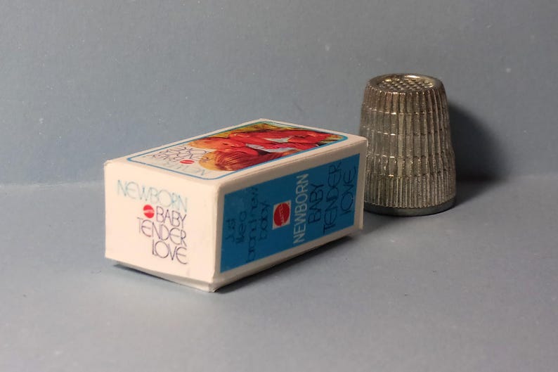 May include: A small white box with the words "NEWBORN BABY TENDER LOVE" in a script font, with a blue border and a picture of a baby on top. A silver thimble is next to the box.