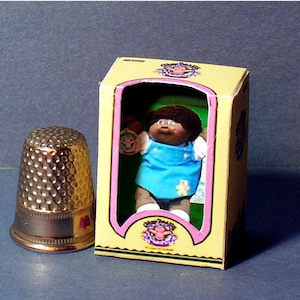 May include: A miniature Cabbage Patch Kid doll in a yellow box with a pink and green border. The doll is wearing a blue dress with a white bib and a white headband. The doll has brown hair and brown eyes.
