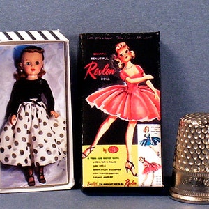 May include: Two boxes of vintage Revlon dolls. One box features a doll in a black top and white polka dot skirt. The other box features a doll in a red dress. The boxes are on a blue surface with a silver thimble.