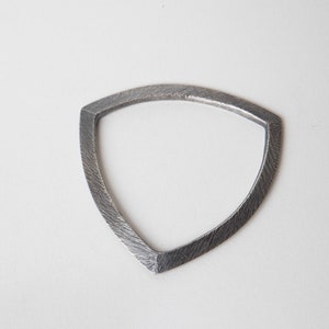 May include: A silver-coloured, shield-shaped metal object with a textured surface. The object has a triangular form with rounded sides and a pointed bottom. The background is plain white.