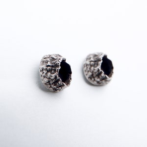 Puede incluir: Dos pequeños objetos redondos, huecos, texturizados y plateados con un interior oscuro. Los objetos son similares en forma y tamaño y están dispuestos uno al lado del otro sobre un fondo blanco.