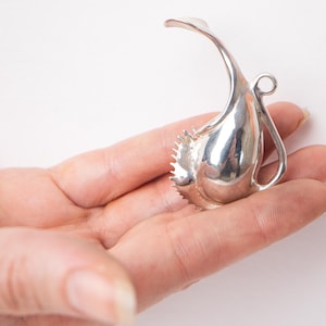 Puede incluir: Un colgante de plata con forma de pájaro estilizado, con un cuerpo texturizado y un bucle para sujetar una cadena.