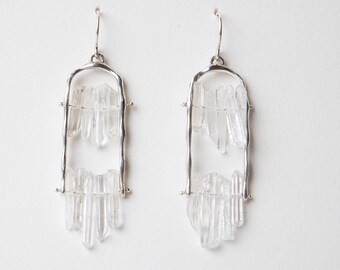 May include: A pair of silver-toned dangle earrings. Each earring features a rectangular frame with clear crystal clusters suspended within. The crystals are of varying lengths, creating a unique, natural aesthetic. The earrings hang from simple, curved ear wires.