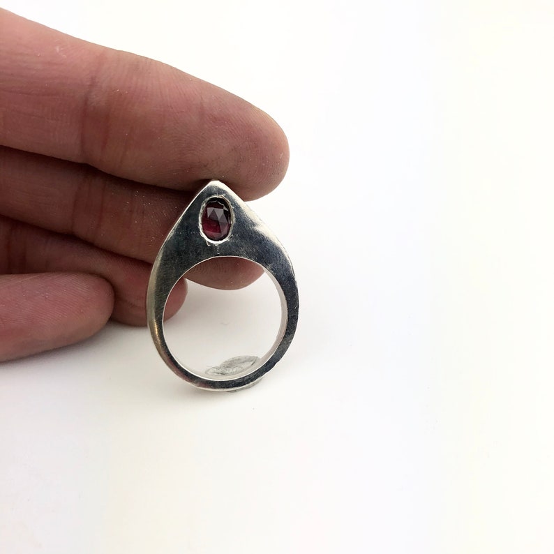 May include: A silver ring with a teardrop-shaped band and a red gemstone set in the top.