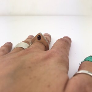 May include: A silver ring with a pointed design and a dark red gemstone set in the center. The ring is worn on a person's finger.