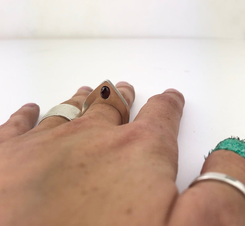 May include: A silver ring with a pointed design and a dark red gemstone set in the center. The ring is worn on a person's finger.