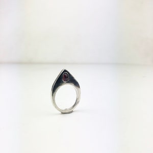 May include: A silver ring with a pointed top and a small, dark red gemstone set in the center.