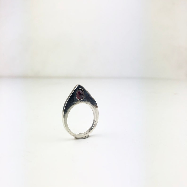 May include: A silver ring with a pointed top and a small, dark red gemstone set in the center.