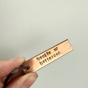 May include: A copper-coloured keychain with a metal ring. The keychain has the words "buckle up buttercup" engraved on it.
