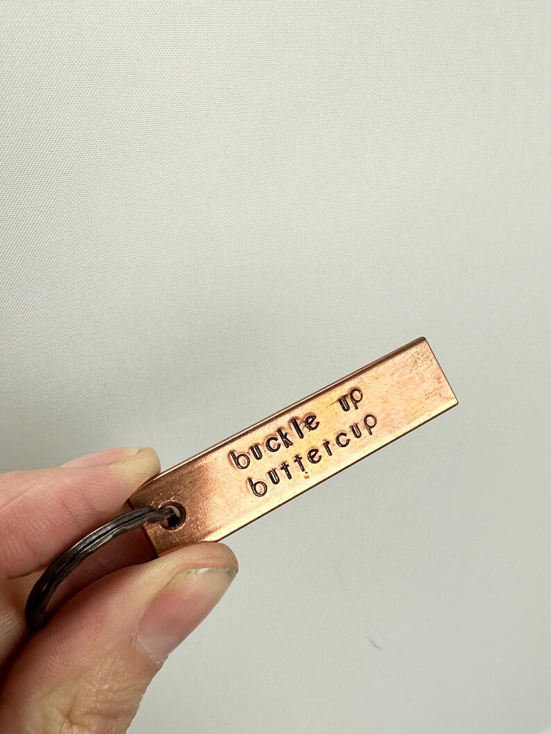 May include: A copper-coloured keychain with a metal ring. The keychain has the words "buckle up buttercup" engraved on it.
